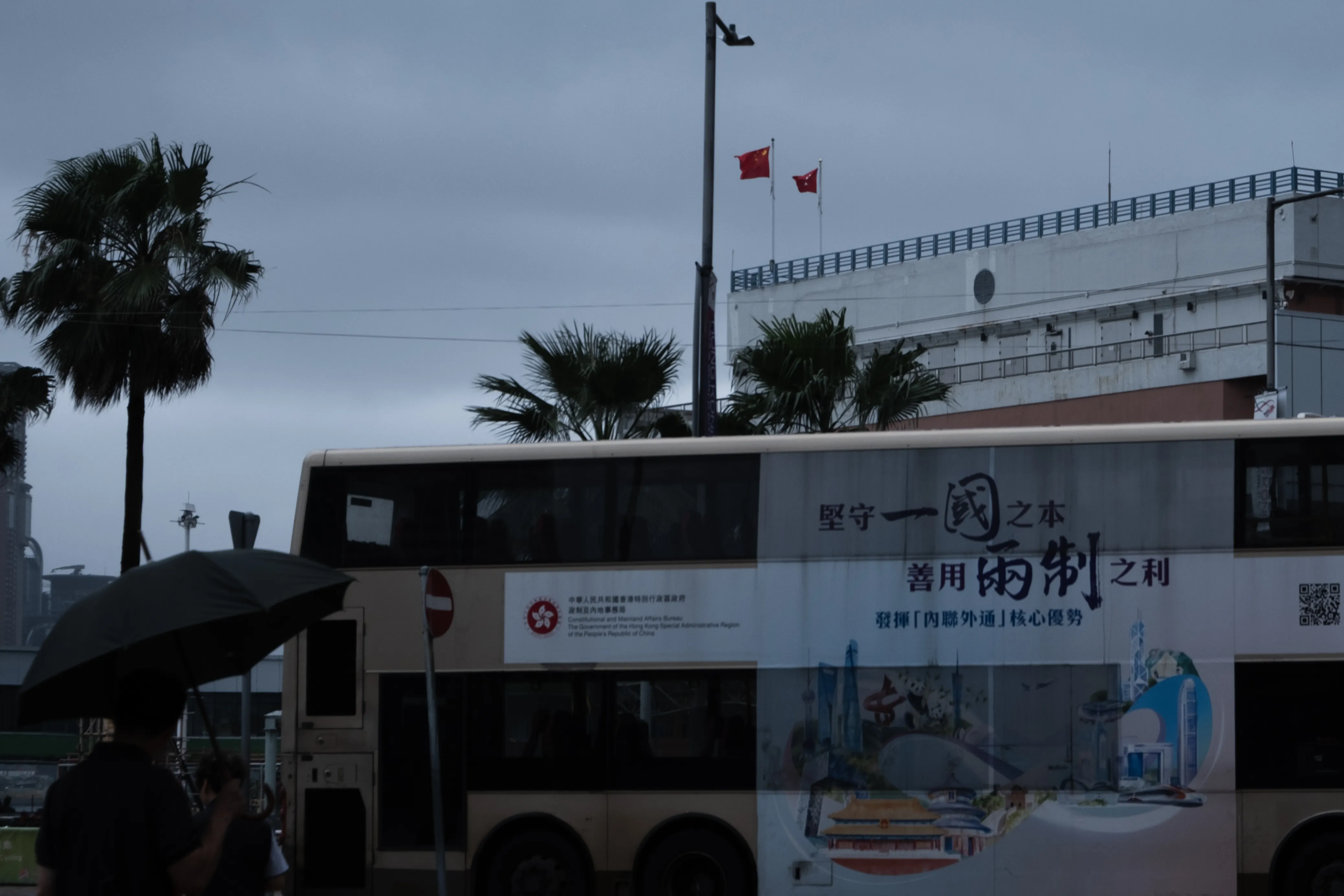 A double-decker bus wrapped in a government advertisement passes under grey skies, with the Chinese and Hong Kong flags on a building behind and a figure holding an umbrella in the foreground