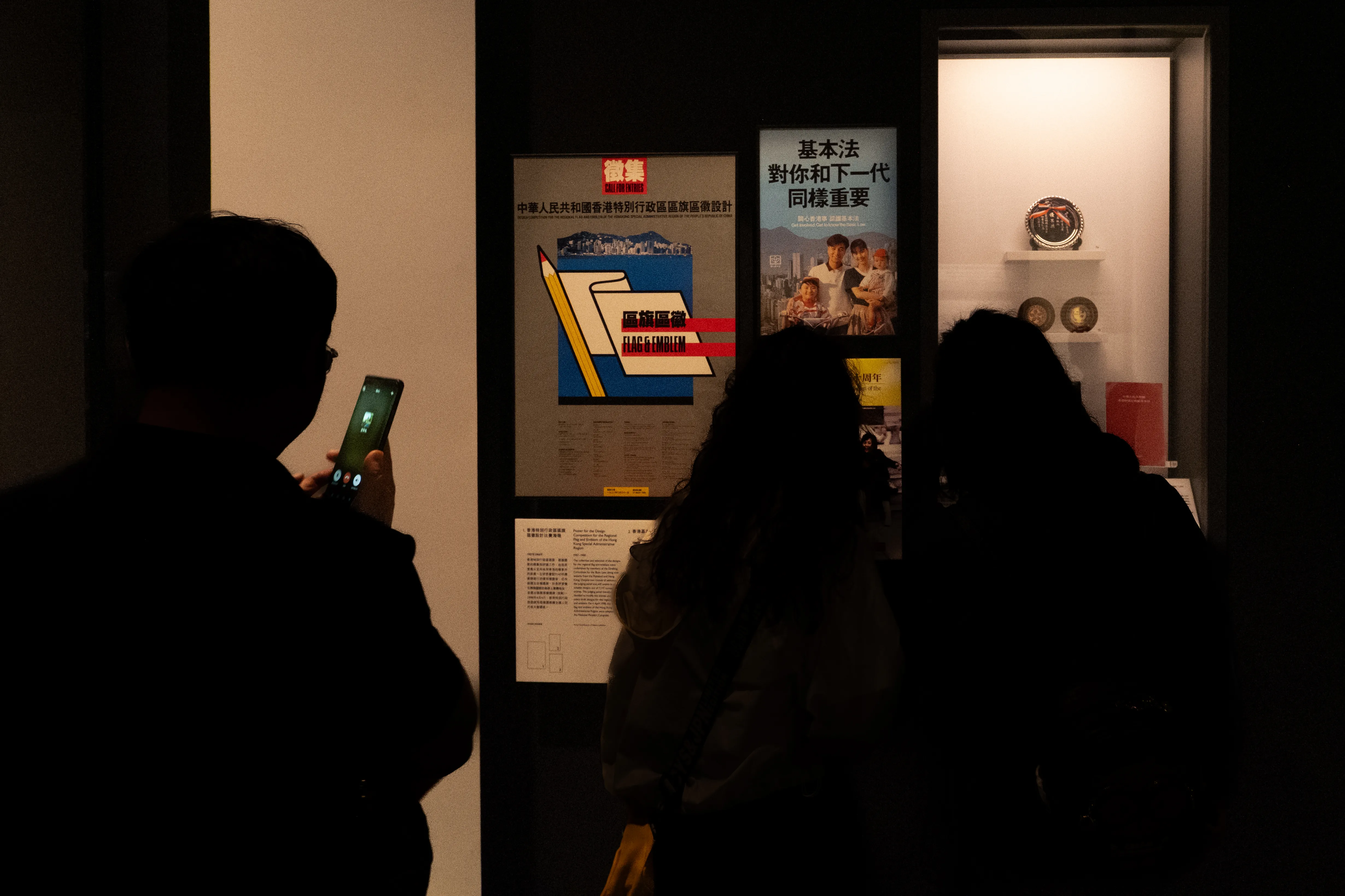 Visitors in silhouette at a dim museum exhibit showing posters from the Hong Kong flag and emblem design competition and a Basic Law poster, one person photographing it with a phone