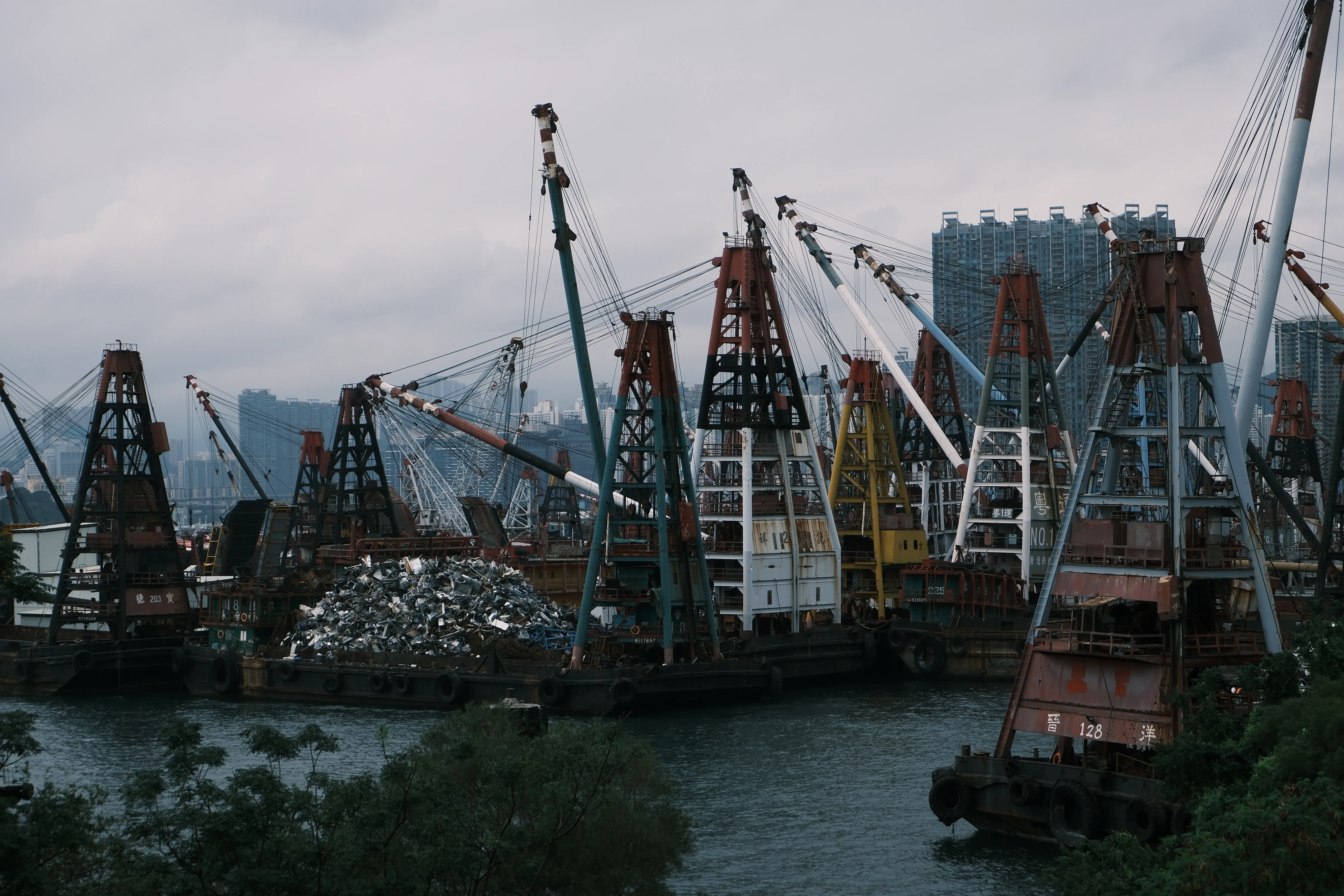 Rows of rusting crane barges and a pile of scrap metal moored in dark water, with high-rise housing blocks behind under an overcast sky