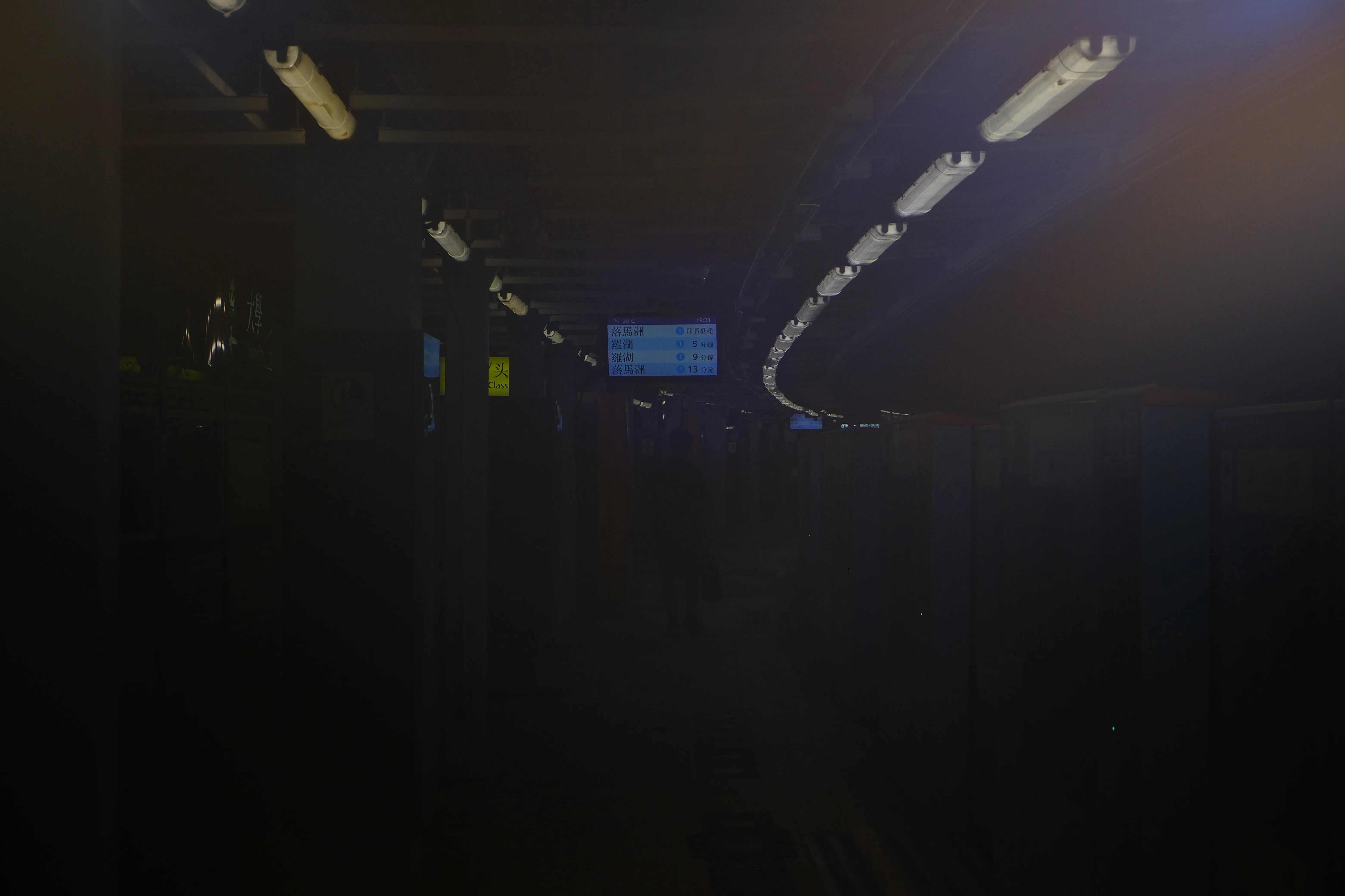 A nearly dark station platform, lit only by a row of ceiling lights and a glowing arrivals screen in the distance