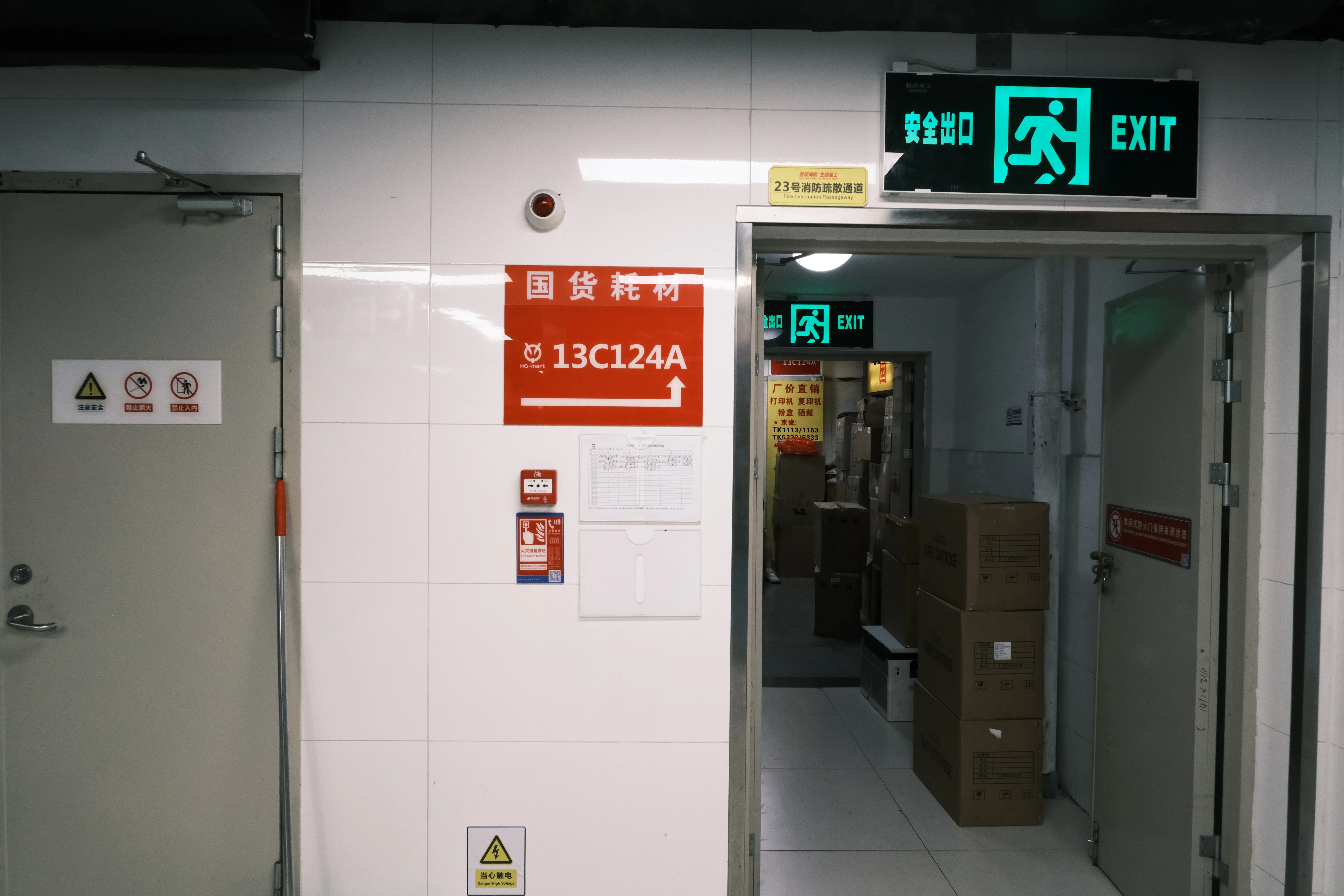A white-tiled back corridor with a green exit sign, a red stall sign reading 国货耗材 13C124A, and cardboard boxes stacked in the next room