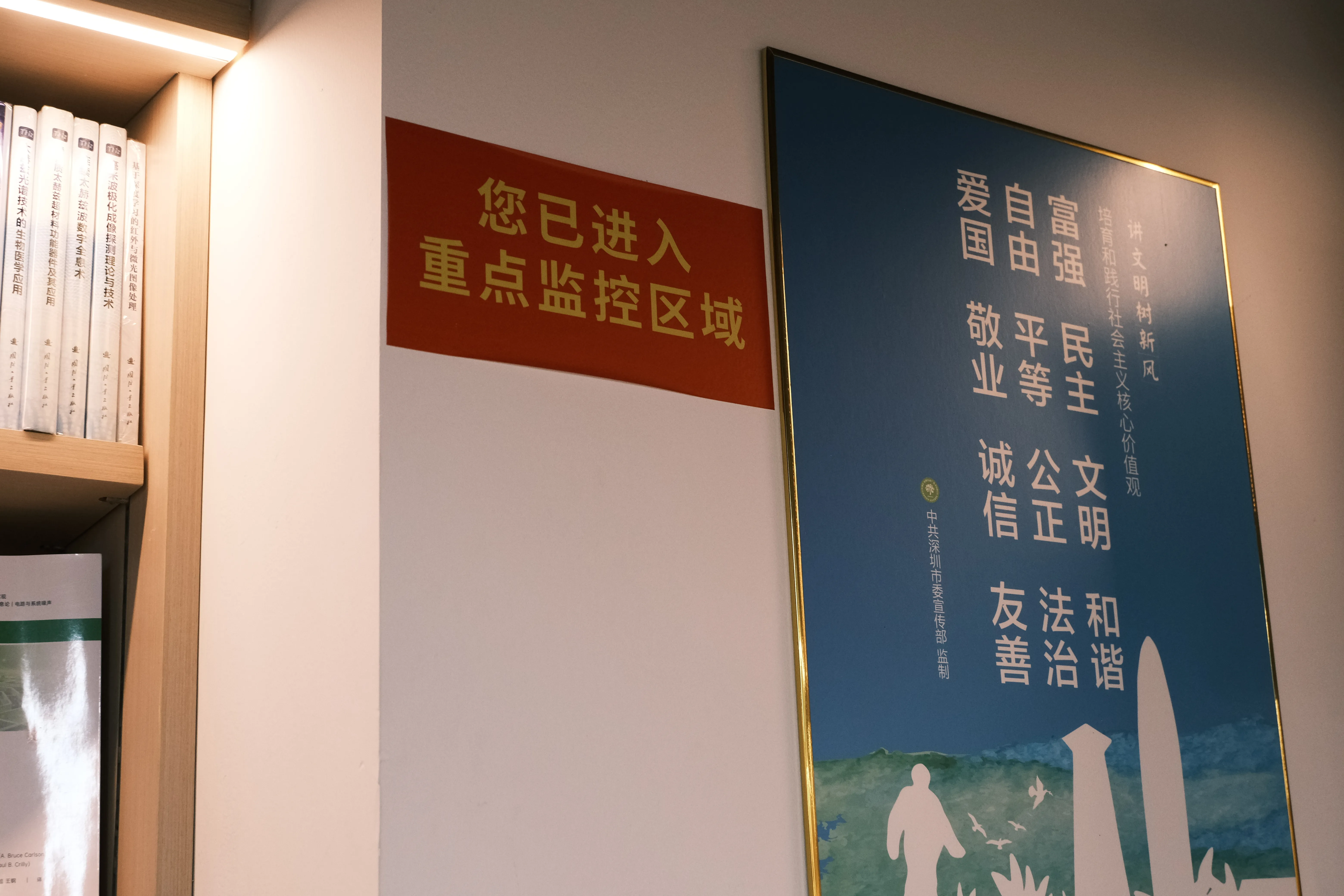 A red sign on a bookstore wall reading 您已进入重点监控区域, next to a framed poster of the Core Socialist Values